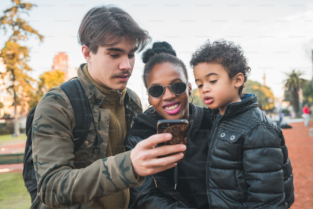 Retrato de una encantadora familia étnica de raza mixta divirtiéndose, relajándose y usando el teléfono móvil en el parque al aire libre.