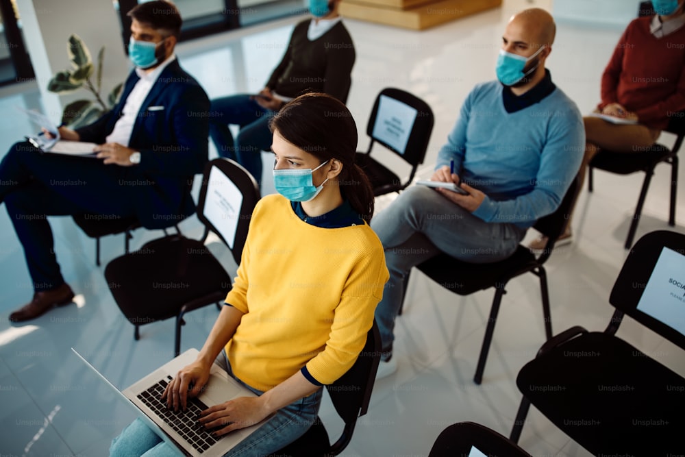 Vista ad angolo alto della donna d'affari che indossa la maschera facciale e utilizza il laptop mentre partecipa a un seminario con i suoi colleghi.