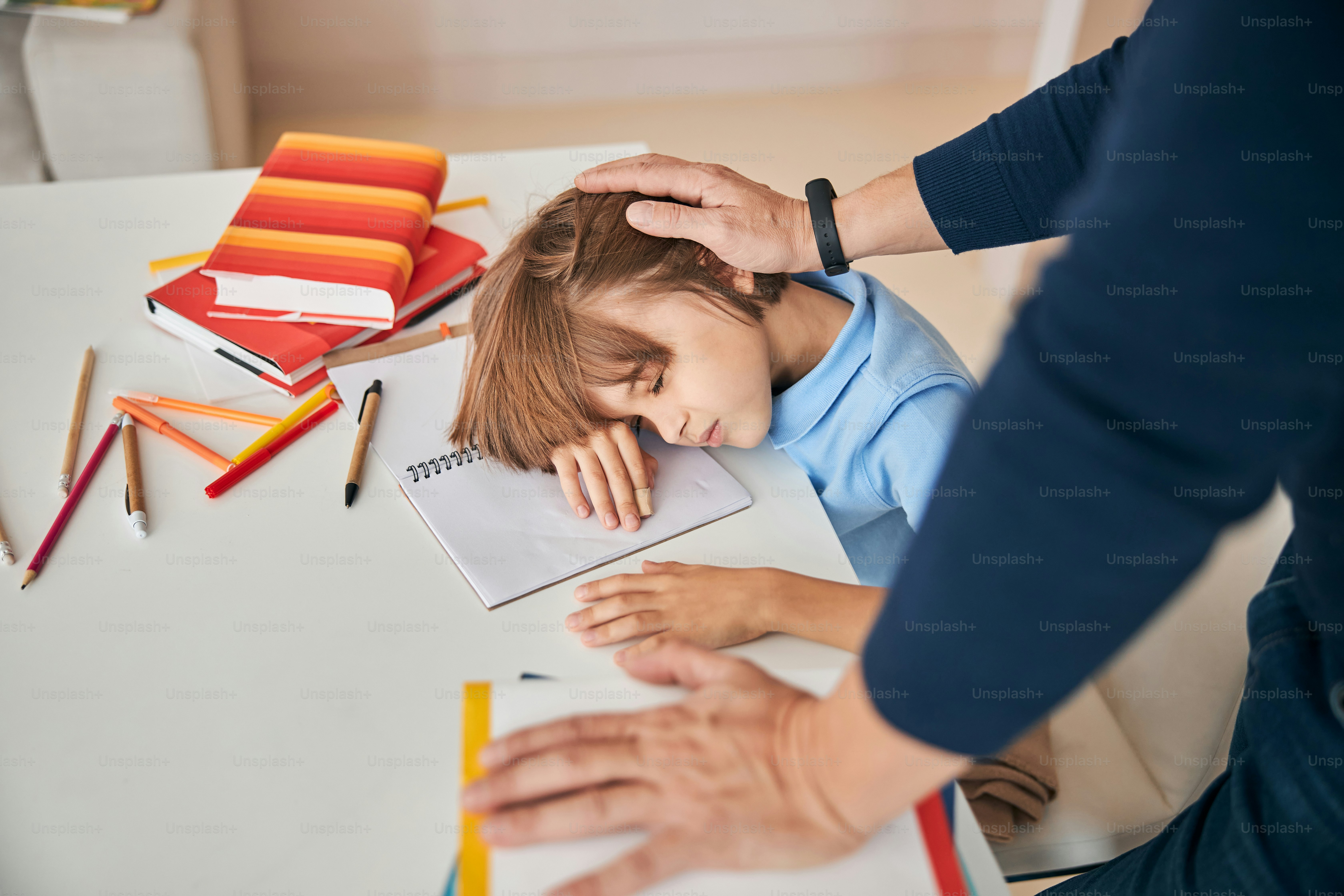 Cute boy taking a nap on his books at the table and unrecognized man patting him on his head