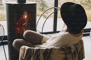 Stylish woman in knitted sweater and hat warming up at modern black fireplace with view on mountains. Cozy warm moments at cold season. Hipster female relaxing in comfortable home