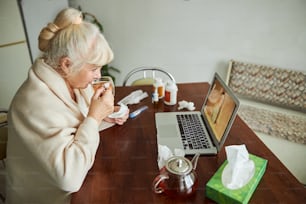 Signora anziana con l'influenza che tiene la tazza di bevanda calda e parla con il medico tramite videochiamata mentre è seduta al tavolo con il computer portatile e i medicamenti
