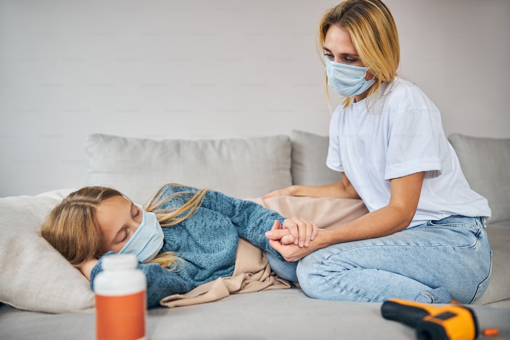 Blonde young Caucasian woman in a face mask sitting next to her sleeping sick daughter