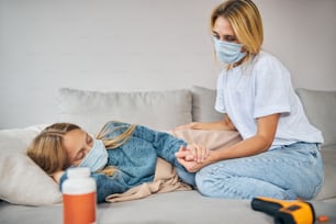 Blonde young Caucasian woman in a face mask sitting next to her sleeping sick daughter