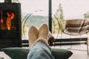 モダンな黒い暖炉と山々の景色を望む大きな窓を背景に、暖かいニットのウールの靴下の足。快適な家でリラックスする女性、居心地の良い暖かい瞬間