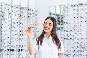 Attraente giovane medico femminile in clinica oftalmologica. Il medico oftalmologo è in piedi vicino agli scaffali con gli occhiali in mano.