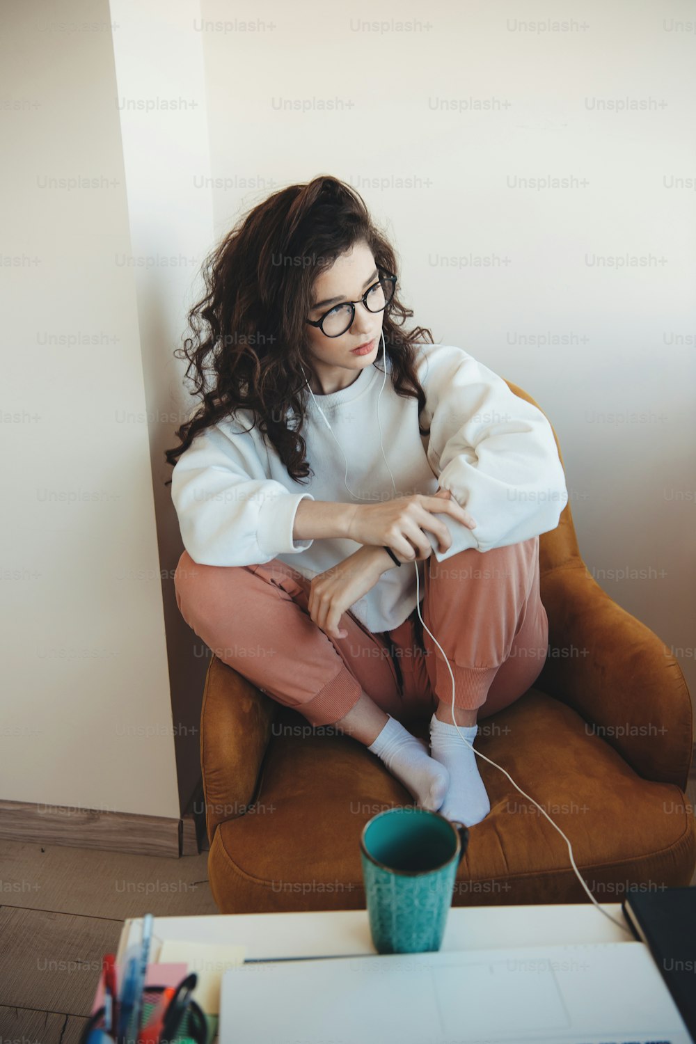 곱슬머리와 안경을 쓴 집중된 백인 여성이 안락의자에 앉아 커피를 마시면서 음악을 듣고 있다