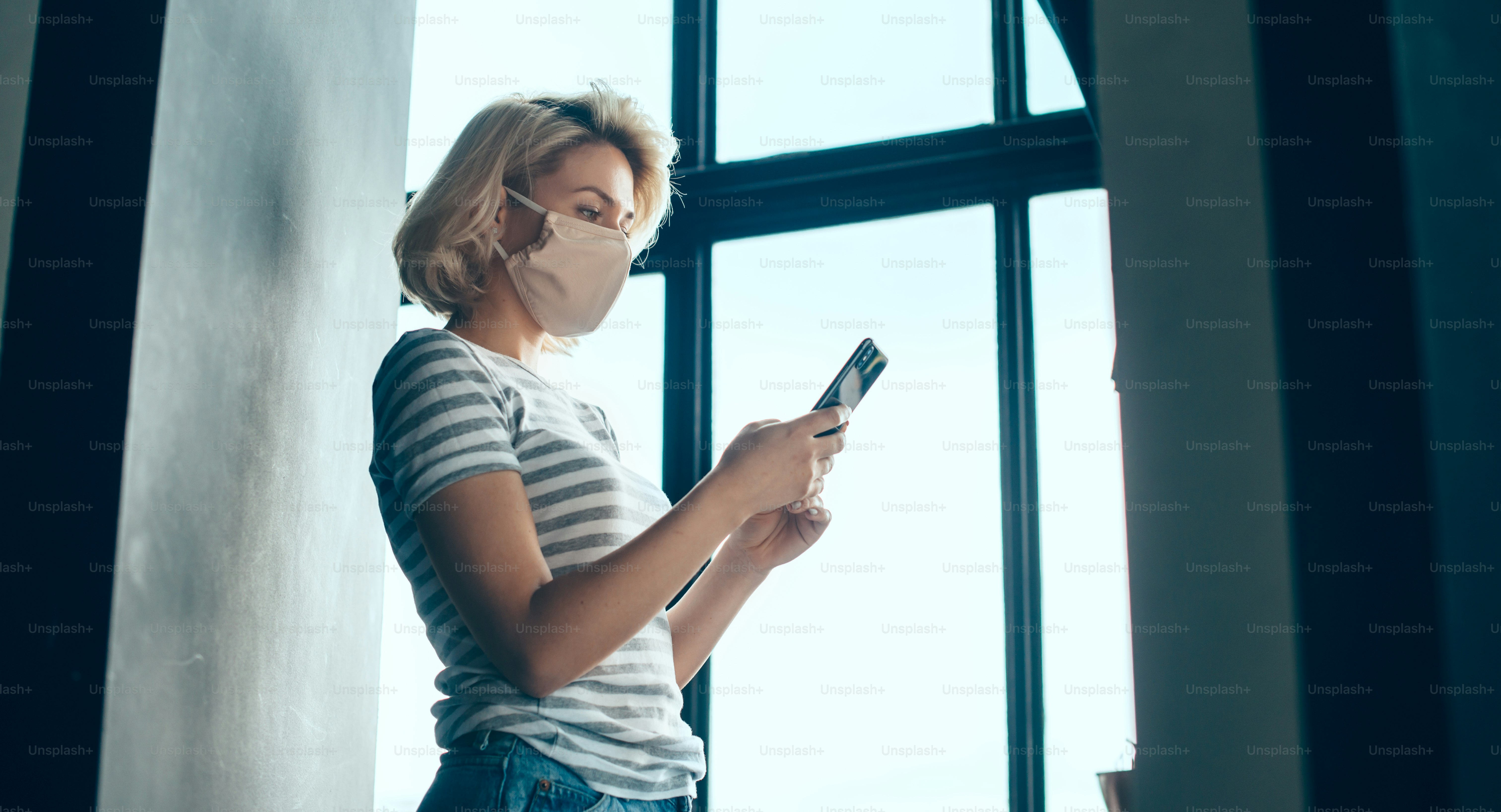 Busy caucasian woman with blonde hair is chatting on phone while wear a mask near the window