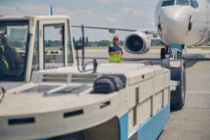 Experienced airport male worker in noise-canceling headphones standing between an airliner and a pushback tractor