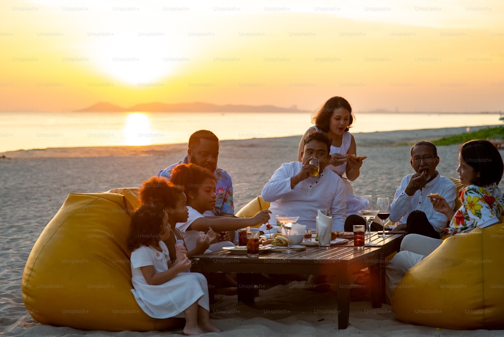 多民族の大家族のグループは、夕暮れ時にビーチで一緒にディナーパーティーを楽しんでいます。
