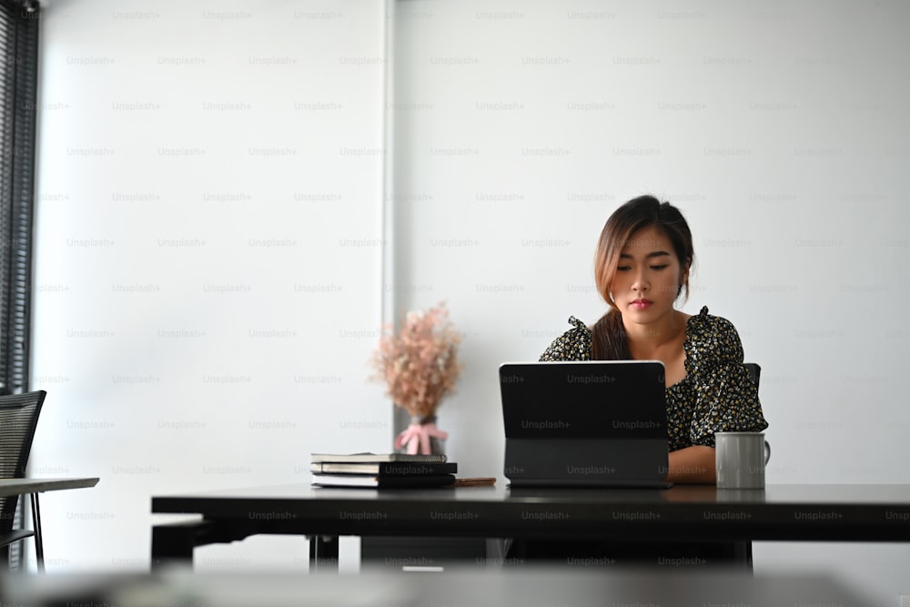 Atractiva joven mujer de negocios segura de sí misma sentada en la mesa de la oficina y trabajando en la tableta.