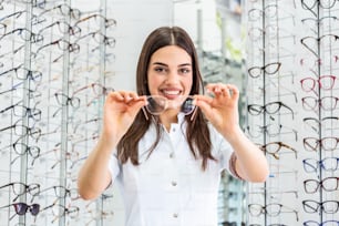 Female optometrist, optician is standing with many glasses in background in optical shop. Stand with spectacles. Eyesight correction. Woman holds spectacles in hands. Closeup.