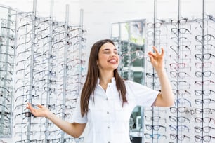 Posing optometrist woman in eyeglasses store smiling looking at camera. Portrait of young smiling woman standing in front of retail display in optical shop