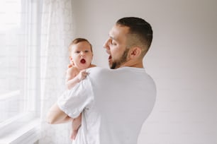 Happy Caucasian father holding newborn baby. Male bearded man parent imitating his yawning child daughter son. Authentic lifestyle candid funny moment. Young dad family life concept.