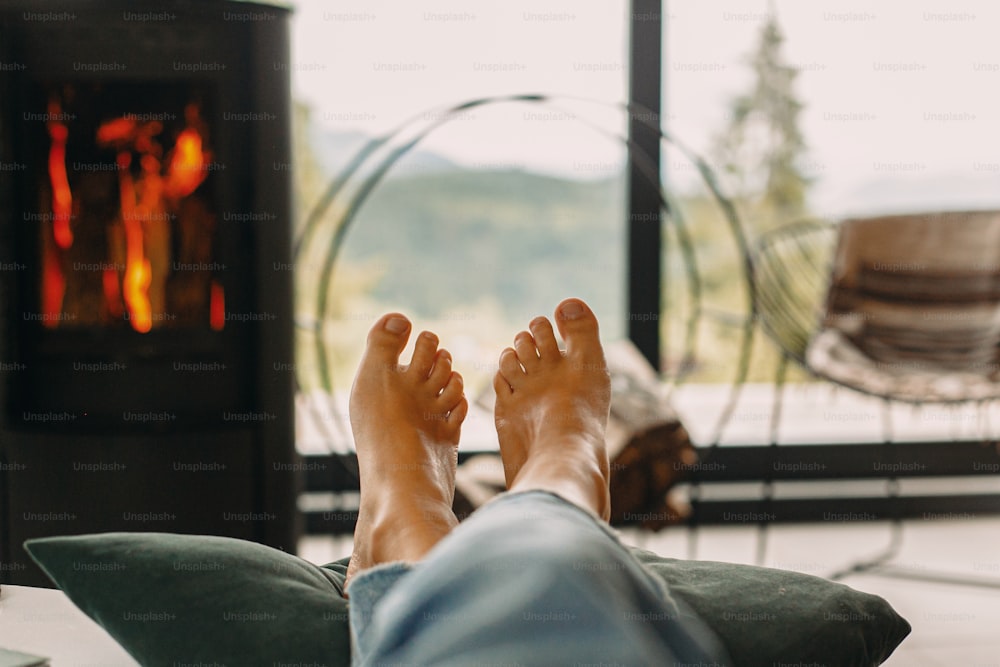 Feet on background of modern black fireplace and big windows with view on mountains. Woman barefoot relaxing in comfortable home, cozy warm moments