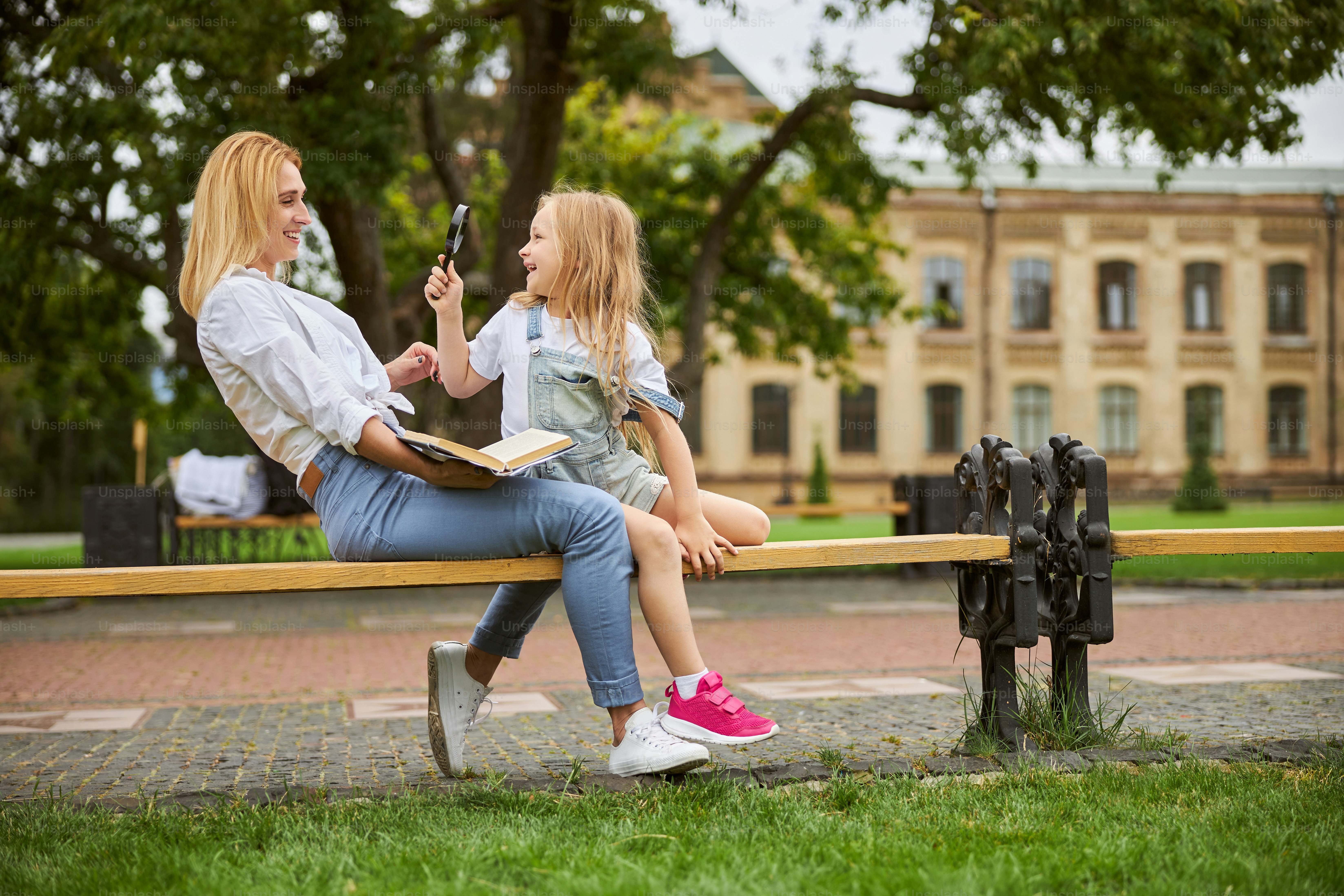 Ganzkörperporträt einer attraktiven Frau, die warmes Wetter im Park genießt, während ein junges weibliches Kind die Glasvergrößerung in der Hand hält