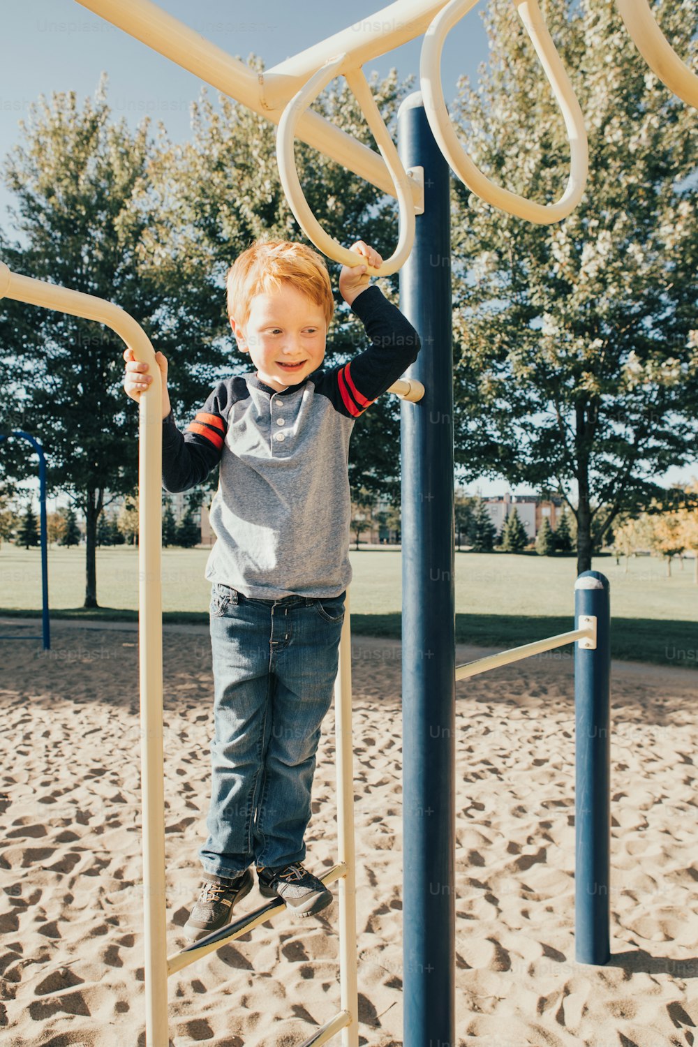 Young red-haired Caucasian boy hanging on monkey bars in park on playground. Summer outdoor activity for kids. Active preschool child doing exercises sport. Healthy happy childhood.