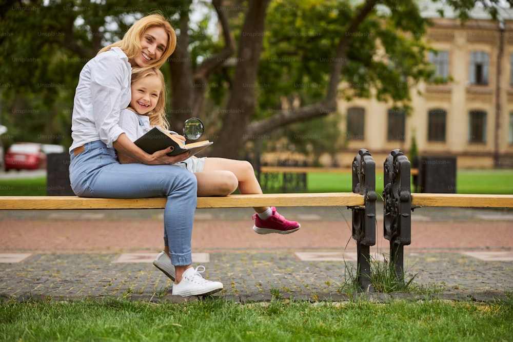Full length portrait of charming female and cute young girl sitting on the wooden bench while reading interesting book in the outdoors