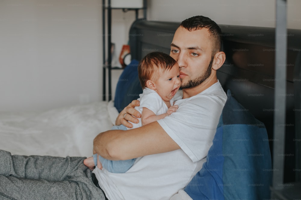 Tired Caucasian father holding newborn yawning baby. Man parent hugging kissing child daughter son in bedroom. Authentic lifestyle candid home moment. Proud young dad. Family fathers day.