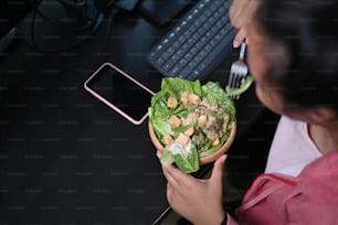 Over head shot of overweight woman eating salad from a bowl at her woprkspace in office.