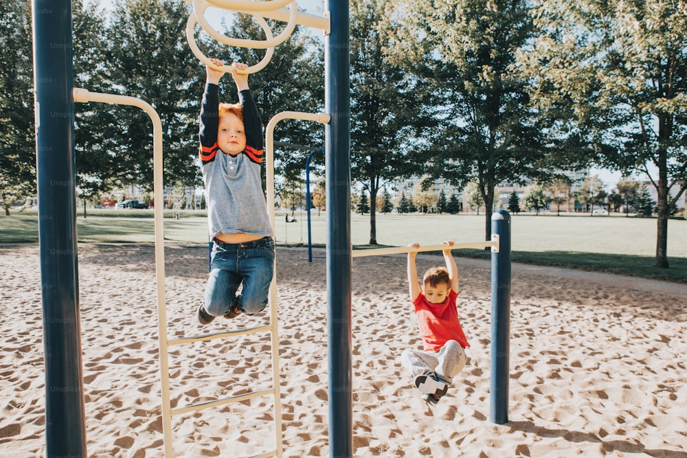 Young Caucasian boys friends hanging on monkey bars and pull-up bars in park on playground. Summer outdoor activity for kids. Active children doing exercises sport. Healthy happy childhood.