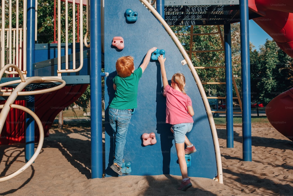 작은 미취학 소년과 소녀는 여름날 놀이터에서 암벽을 등반합니다. 행복한 어린 시절 생활 방식 개념입니다. 아이들을 위한 계절별 야외 활동. 뒤에서 본 모습.
