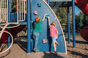 Niño y niña de preescolar escalando la pared de roca en el patio de recreo afuera en el día de verano. Concepto de estilo de vida de la infancia feliz. Actividad al aire libre de temporada para niños. Vista desde atrás.