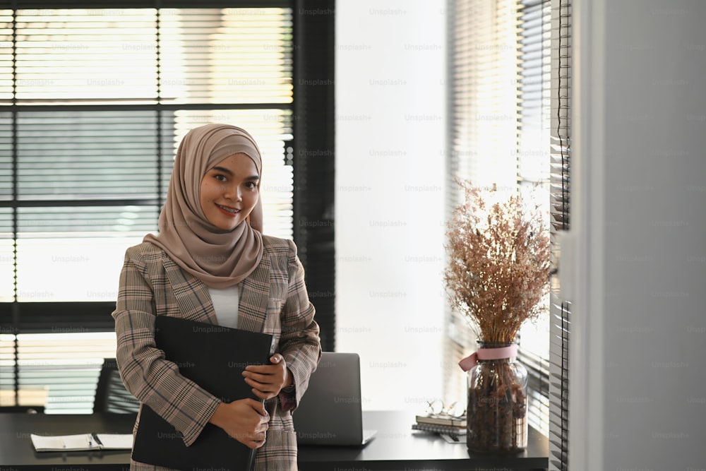 히잡을 쓴 무슬림 사업가의 초상화가 사무실에 서 있는 동안 문서를 들고 카메라를 향해 웃고 있다.