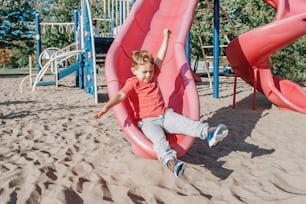 Active happy funny smiling Caucasian boy child sliding on playground schoolyard outdoor on summer sunny day. Kid having fun. Seasonal kids activity outside. Authentic childhood lifestyle concept.