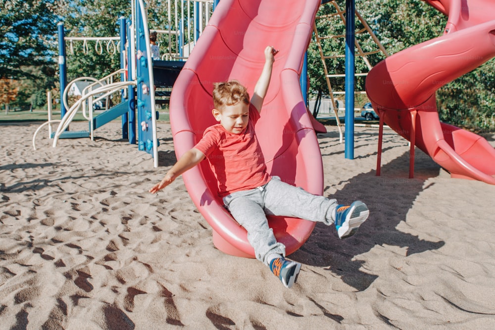 Active happy funny smiling Caucasian boy child sliding on playground schoolyard outdoor on summer sunny day. Kid having fun. Seasonal kids activity outside. Authentic childhood lifestyle concept.
