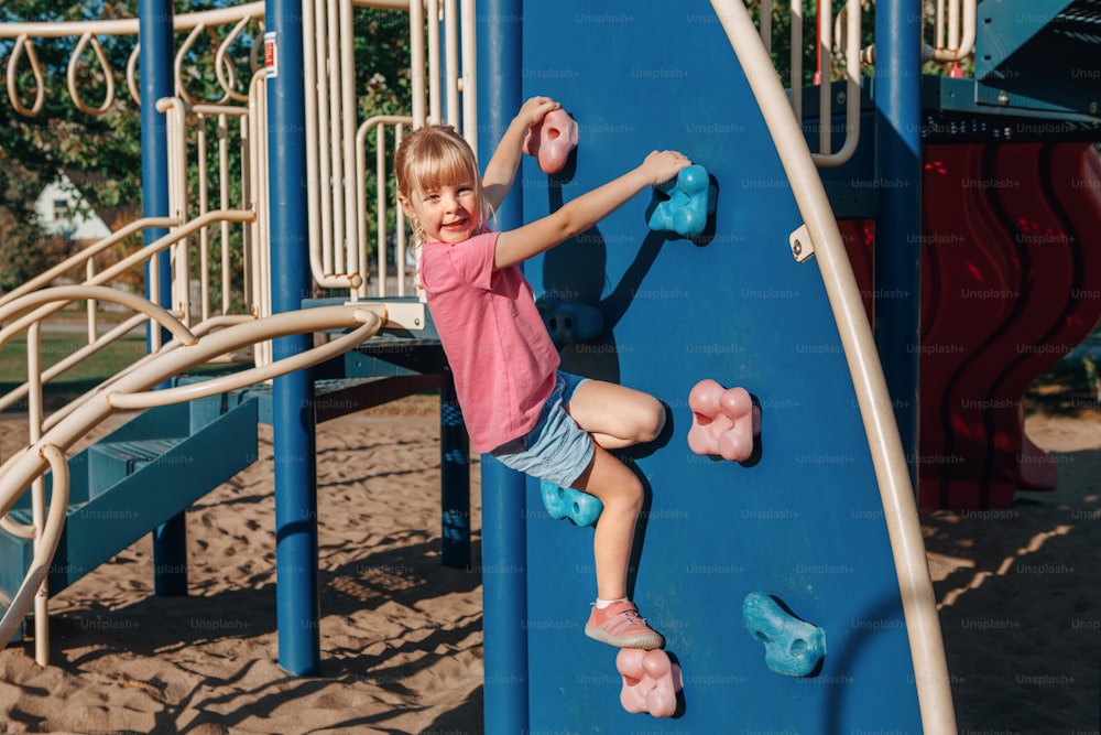Menina pré-escolar sorridente feliz escalando parede de rocha no playground do lado de fora no dia de verão. Conceito de estilo de vida feliz na infância. Atividade sazonal ao ar livre para crianças.