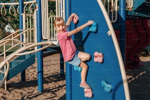 Happy smiling little preschool girl climbing rock wall at playground outside on summer day. Happy childhood lifestyle concept. Seasonal outdoor activity for kids.