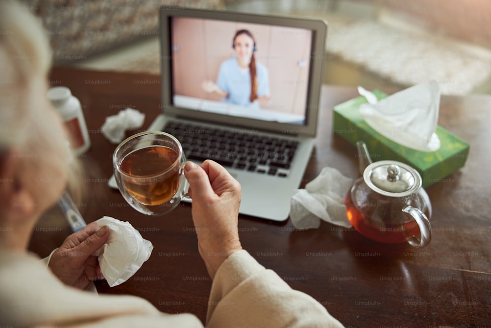 Close up of elderly woman holding cup of hot drink and talking with doctor through video call while sitting at the table with laptop