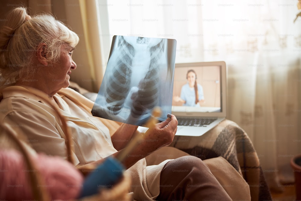 Nette alte Dame, die im Sessel sitzt und Röntgenaufnahmen der Brust hält, während sie mit der Therapeutin per Videoanruf spricht