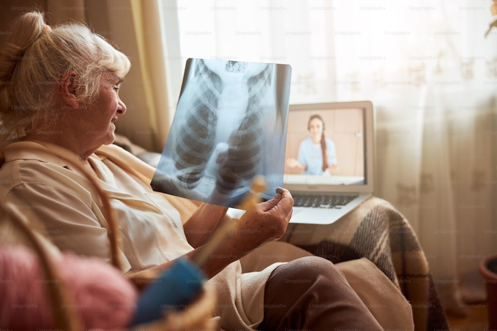 Nice old lady sitting in armchair and holding chest x-ray while talking with female therapist through video call