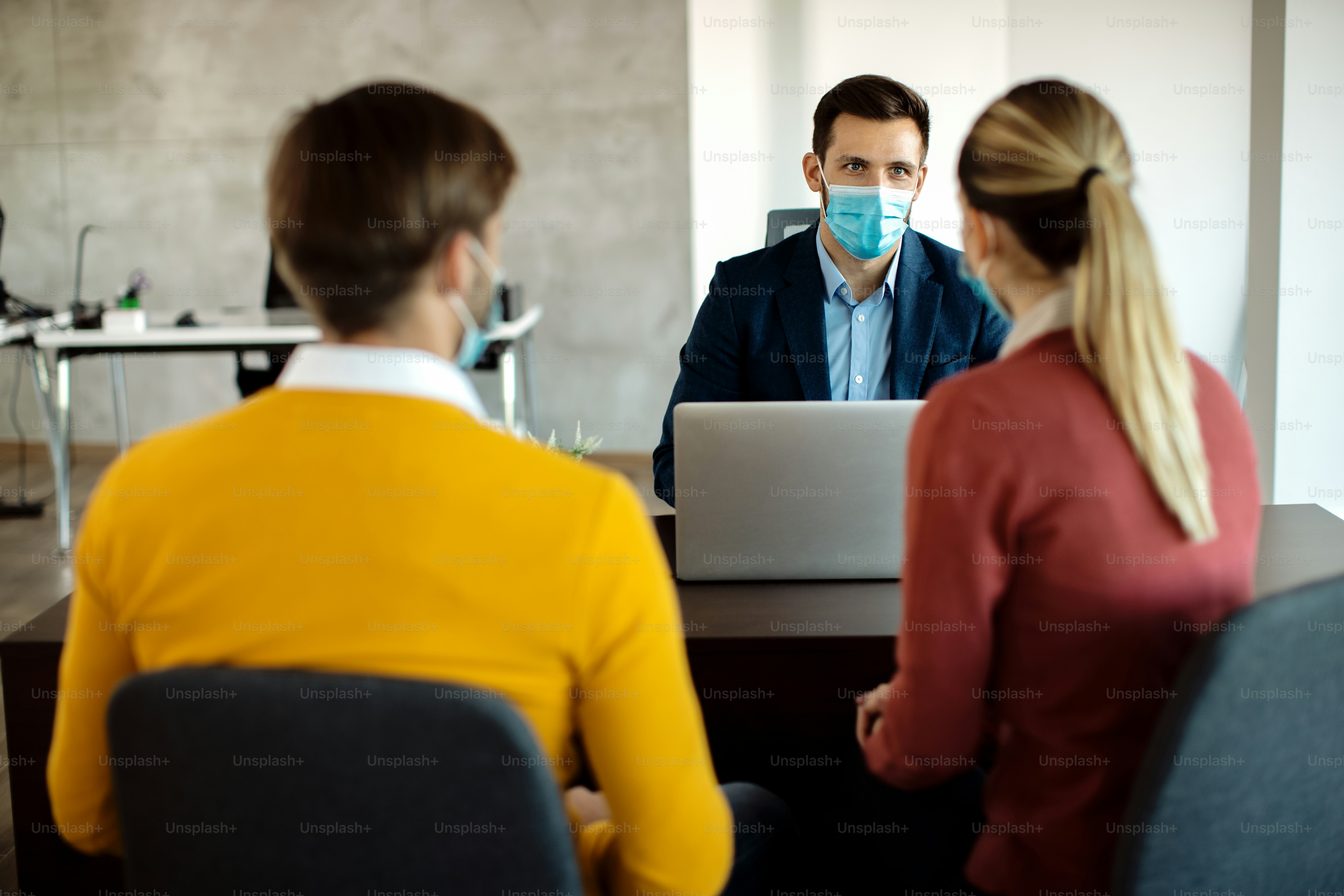 Insurance agent using laptop while communicating with a couple in the office and wearing face mask due to coronavirus pandemic.