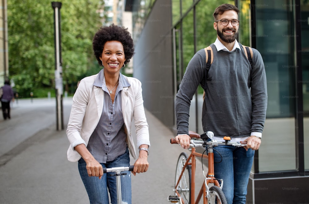 Zwei lächelnde Geschäftsleute fahren Elektroroller, Fahrrad vor modernem Geschäftsgebäude
