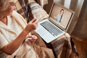 Elderly lady sitting in front of laptop and holding digital thermometer while talking with female doctor through video call
