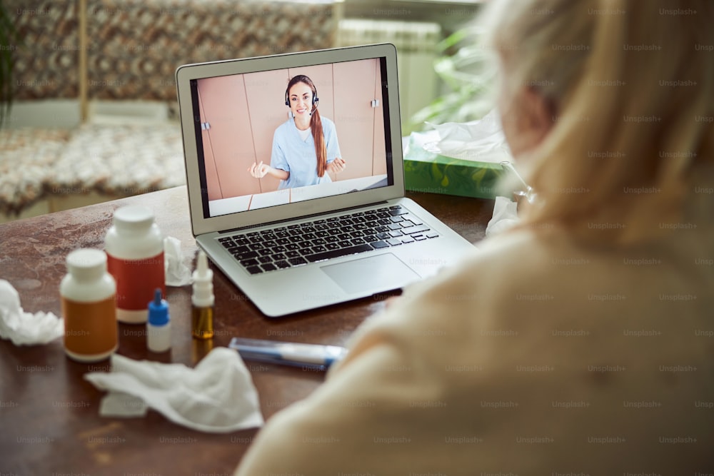 Sick elderly woman sitting at the table with laptop and talking with female doctor through video call