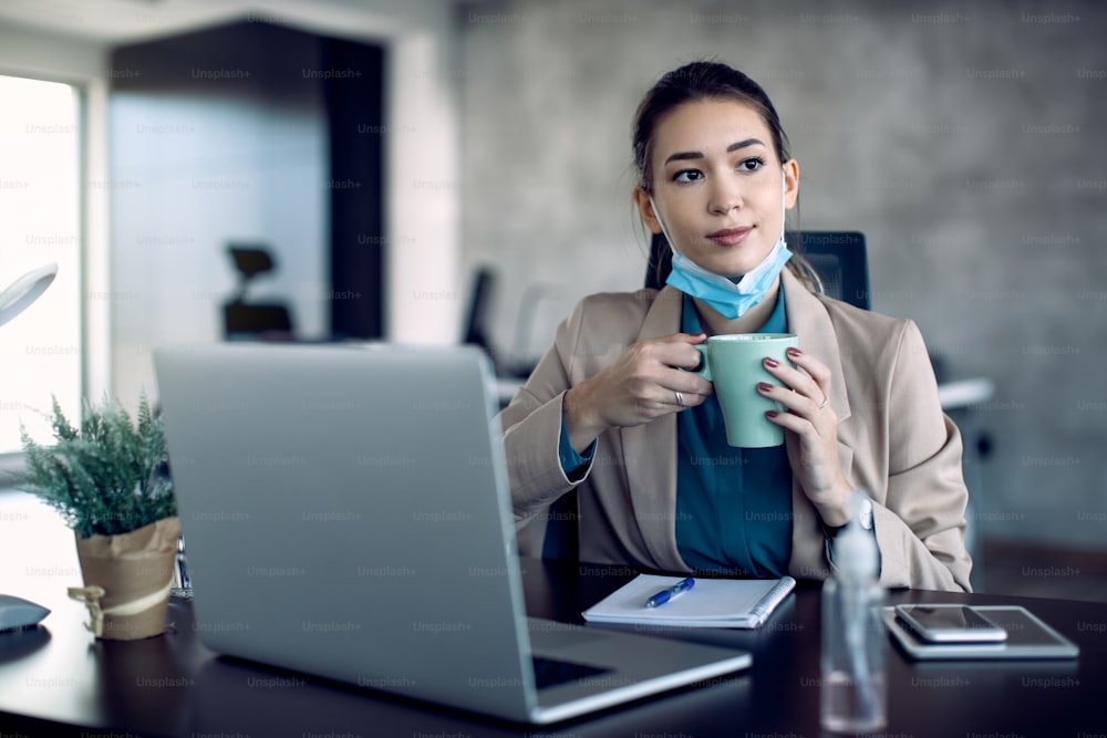 Jeune femme d’affaires prenant une tasse de café et pensant à quelque chose tout en travaillant au bureau pendant la pandémie de coronavirus.