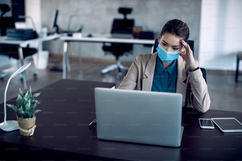 Jeune femme d’affaires s’inquiète en lisant de mauvaises nouvelles sur un ordinateur au bureau. Elle porte un masque facial en raison de la pandémie de COVID-19.