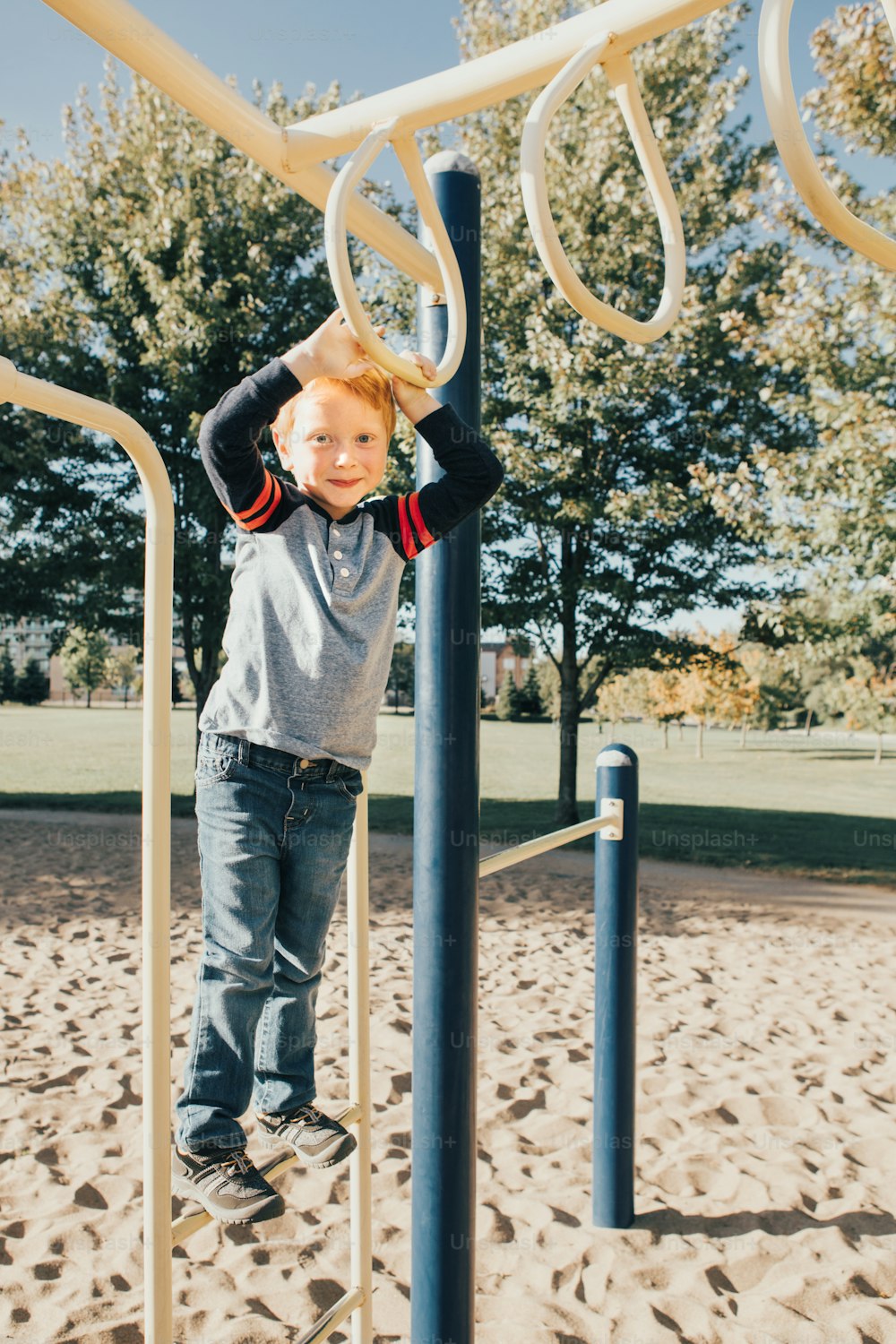 Young Caucasian boy hanging on monkey bars in park on playground. Summer outdoor activity for kids. Active preschool child doing exercises sport. Healthy happy childhood.