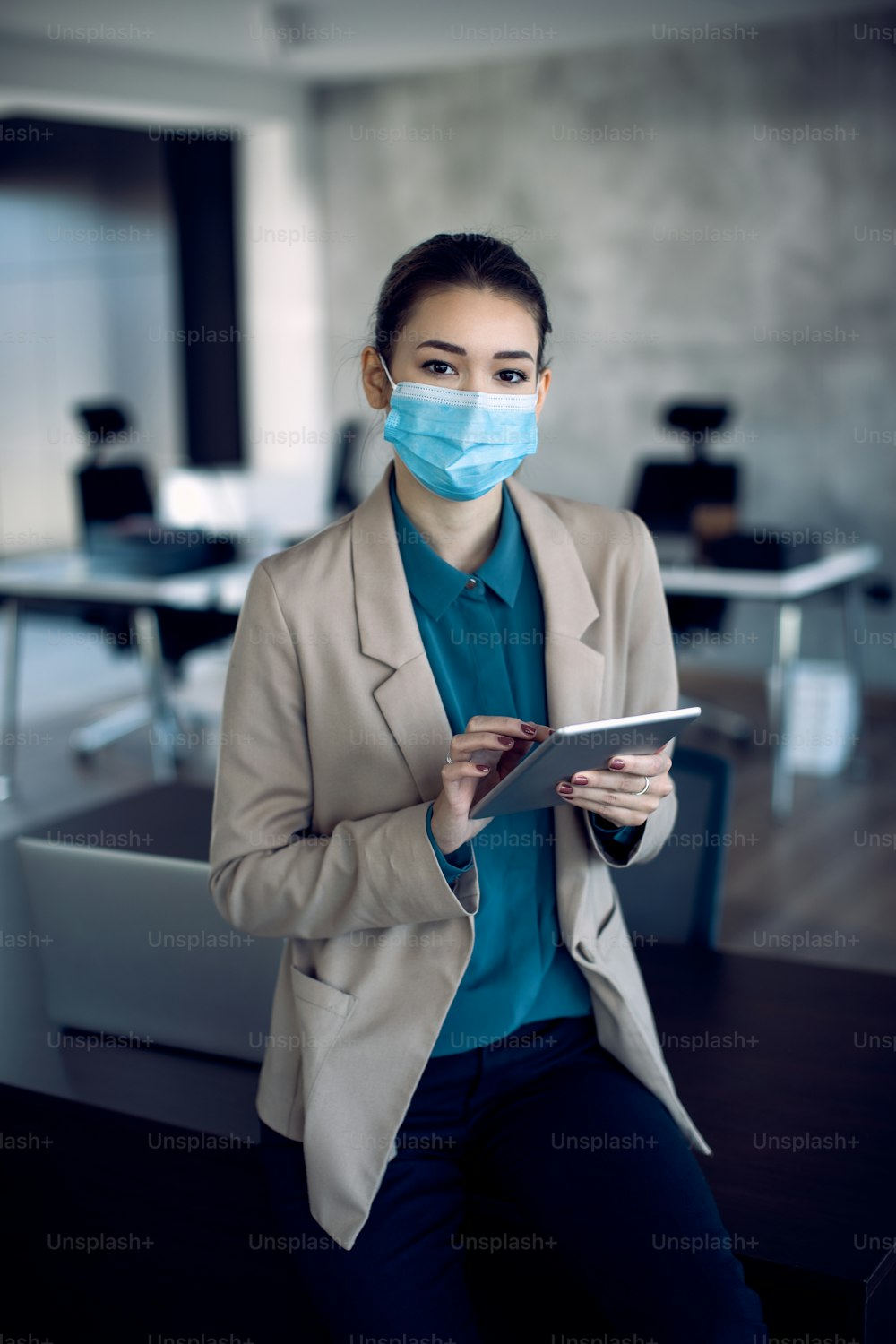 Young businesswoman using touchpad while wearing face mask in the office and looking at camera.