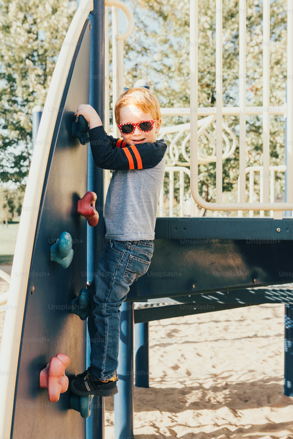 Niño preescolar sonriente feliz con gafas de sol escalando la pared de roca en el patio de recreo afuera en el día de verano. Concepto de estilo de vida de la infancia feliz. Actividad al aire libre de temporada para niños.
