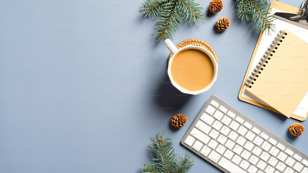 커피 컵, 키보드, 종이 노트북, 솔방울, 파스텔 블루 배경에 가지가 있는 작업 공간. 크리스마스, 겨울 휴가 개념. 아늑하고 휘게, 북유럽 스타일.
