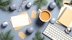 Flat lay mesa de escritório em casa de Natal com ramos de pinho, teclado, xícara de café, bolas, decorações de Natal em fundo azul pastel. Aconchegante, hygge, estilo nórdico.