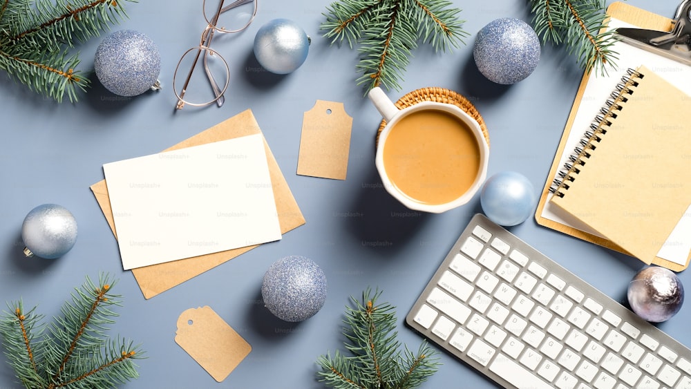 Flat lay Christmas home office desk with pine branches, keyboard, coffee cup, balls, Christmas decorations on pastel blue background. Cozy, hygge, nordic style.