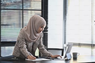 A professional young muslim businesswoman is using tablet computer work and writing work plan on personal notebook.