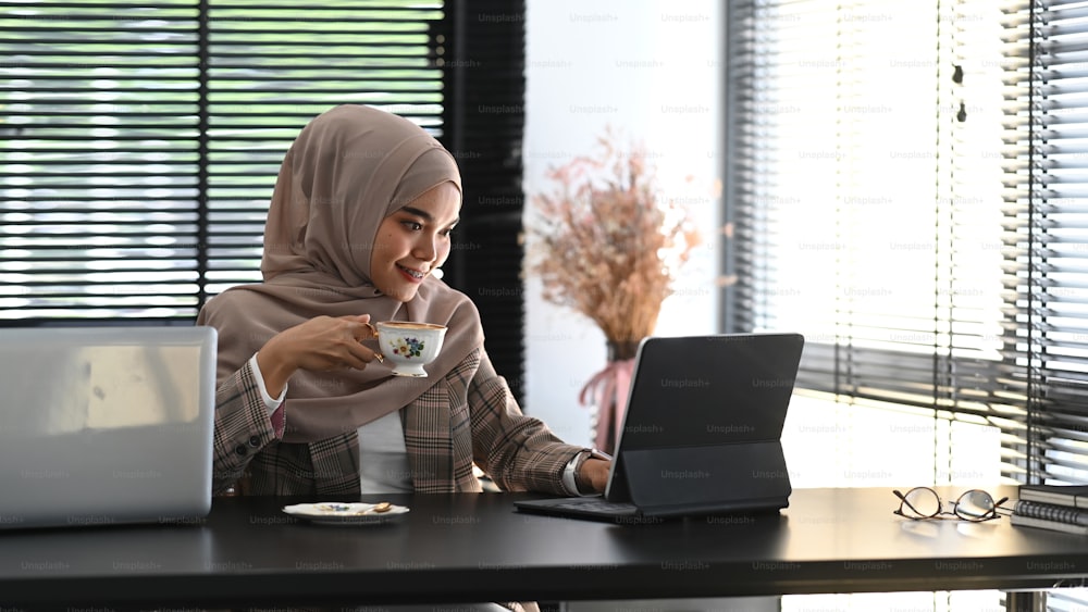 히잡을 쓴 무슬림 사업가가 책상에 앉아 노트북 컴퓨터로 작업하고 커피나 차를 마시고 있다.