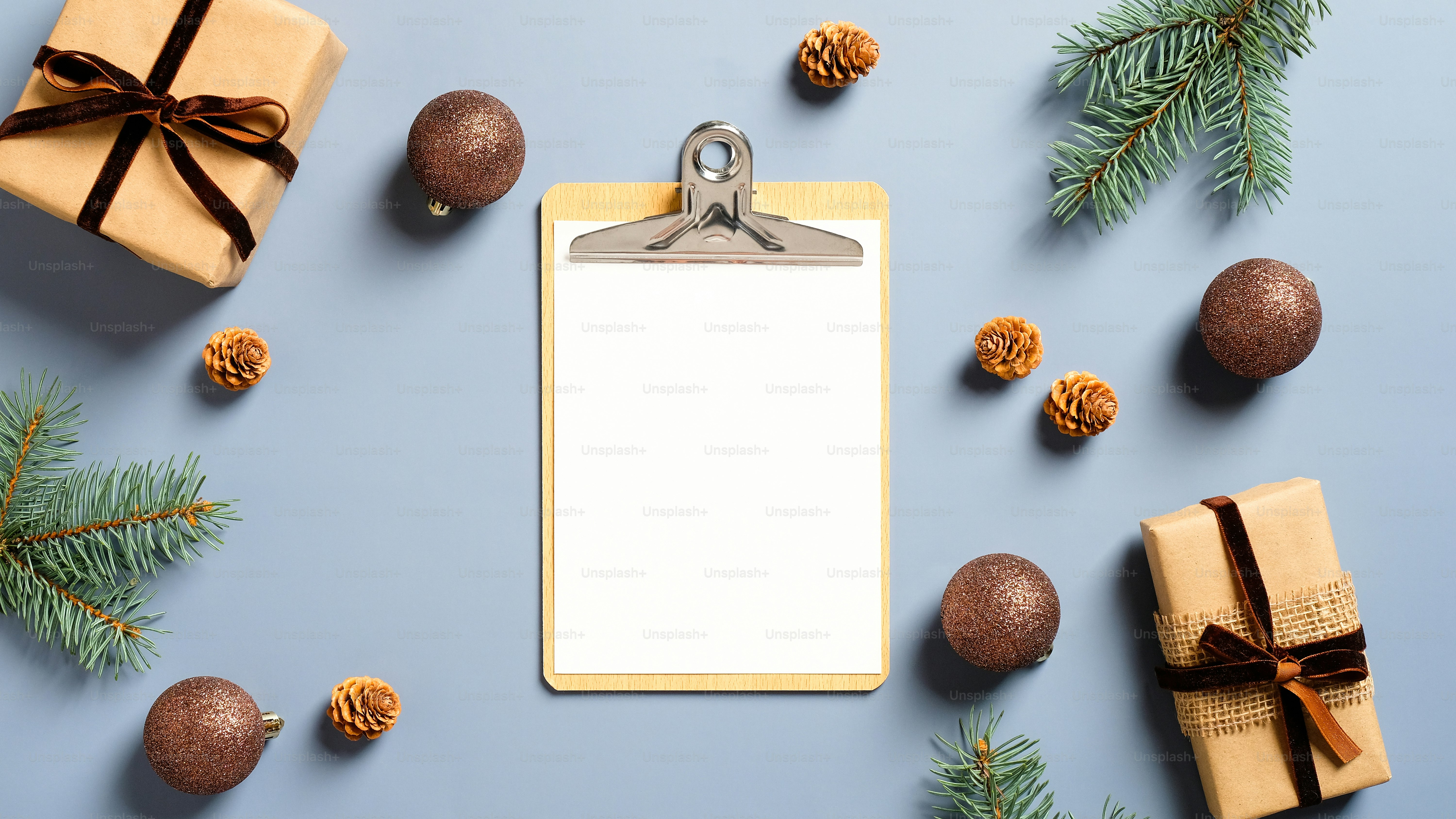 Festive workspace table with blank paper clipboard, gift boxes, fir branches, balls on pastel blue background. Christmas, winter holidays concept. Minimal, nordic style.