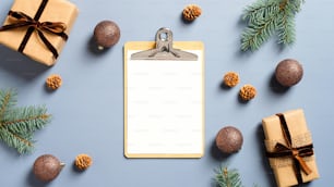 Festive workspace table with blank paper clipboard, gift boxes, fir branches, balls on pastel blue background. Christmas, winter holidays concept. Minimal, nordic style.